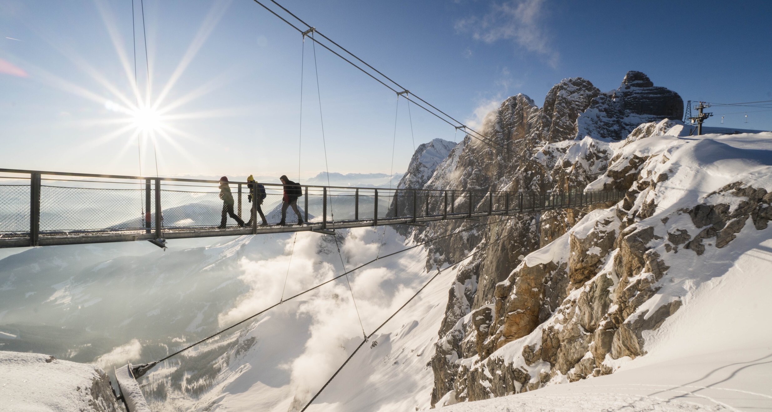 Three hikers cross a snowy suspension bridge high in the mountains