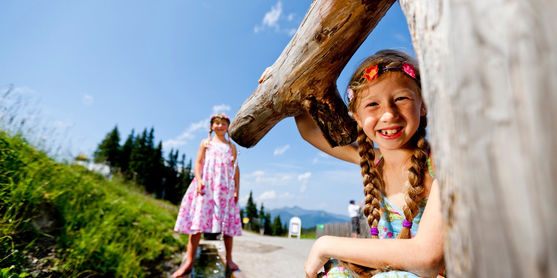 Two girls outdoors, one smiling with braided hair near a large log. | © Tom Lamm