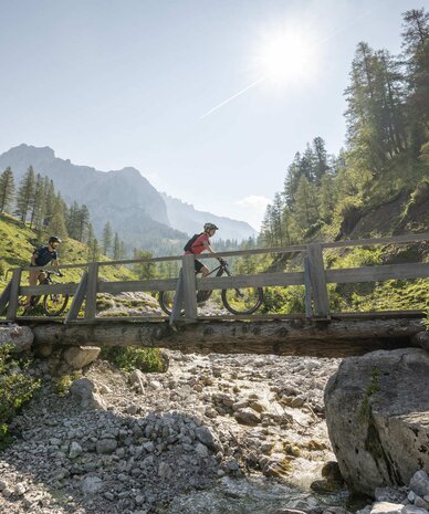 Two cyclists on a wooden bridge over a rocky stream.
