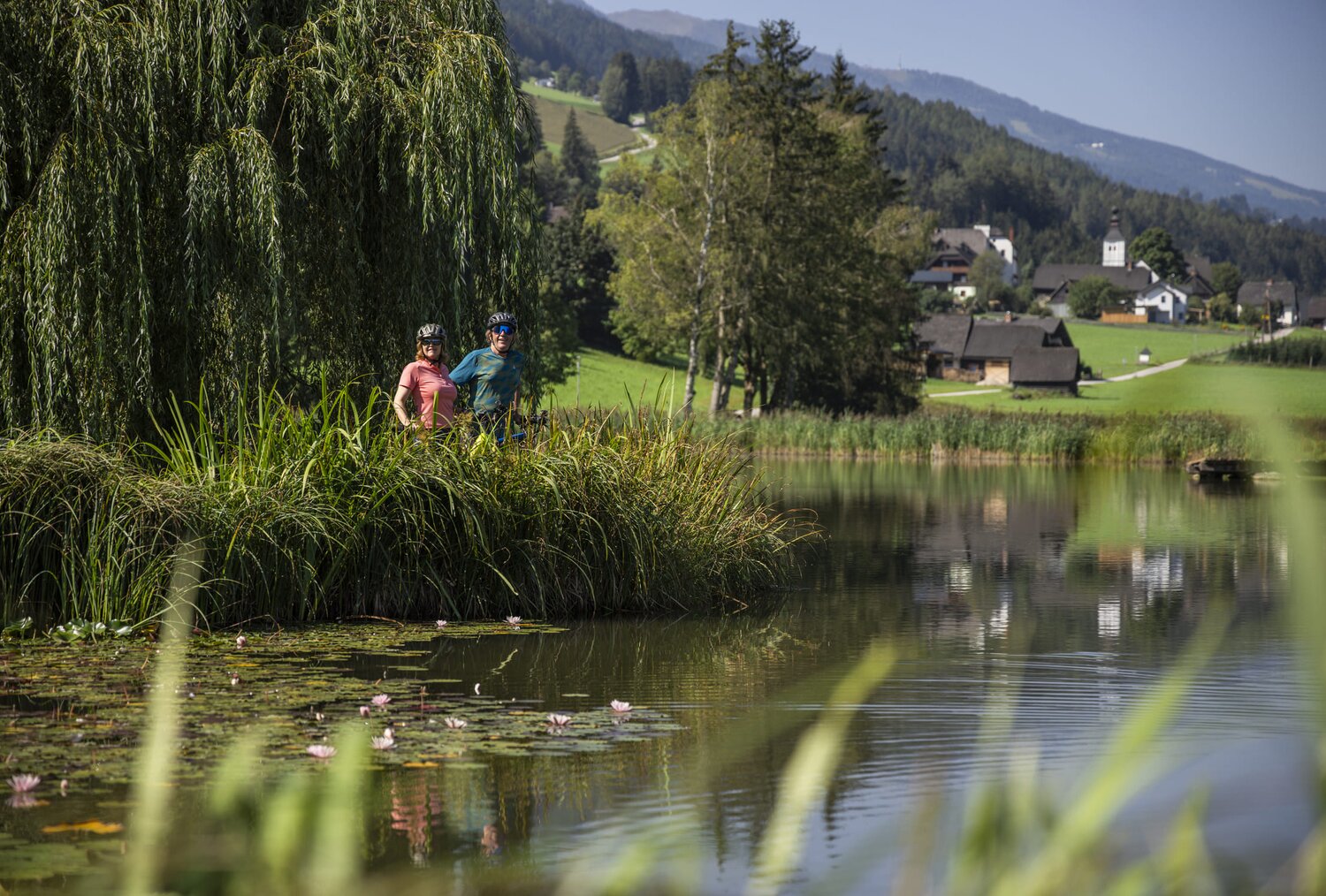 Two cyclists stand at the edge of a pond with reeds.