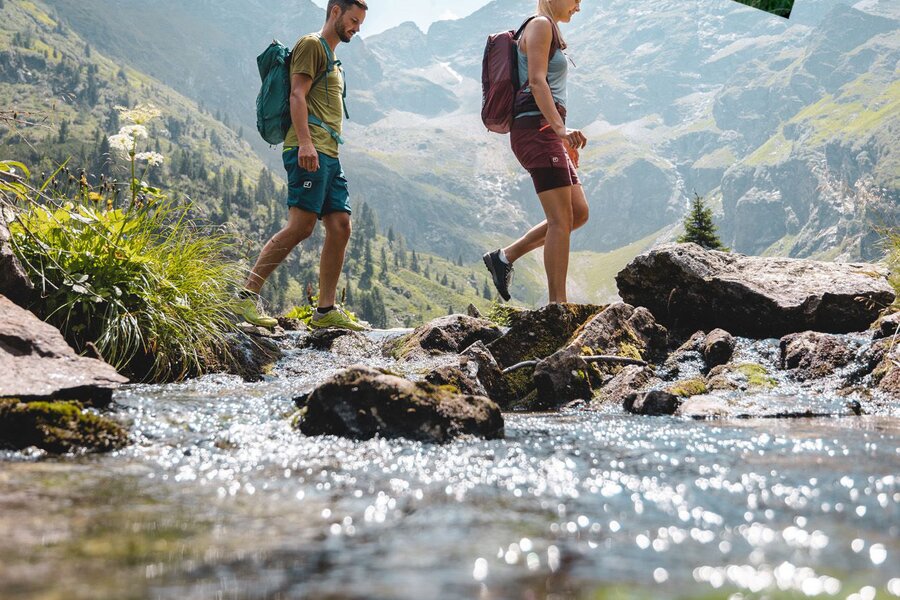 Hikers cross rocky stream beneath mountains; Sommer Card banner.