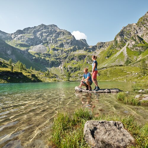 Two hikers stand on a rock in a clear mountain lake