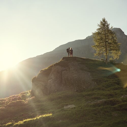 Two hikers stand on a rocky hilltop at sunset with mountains