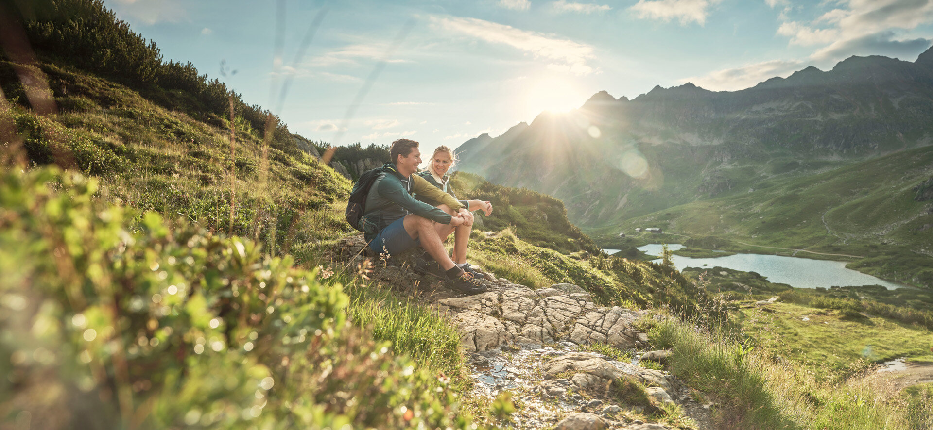 Two hikers rest on a rocky slope overlooking alpine valley