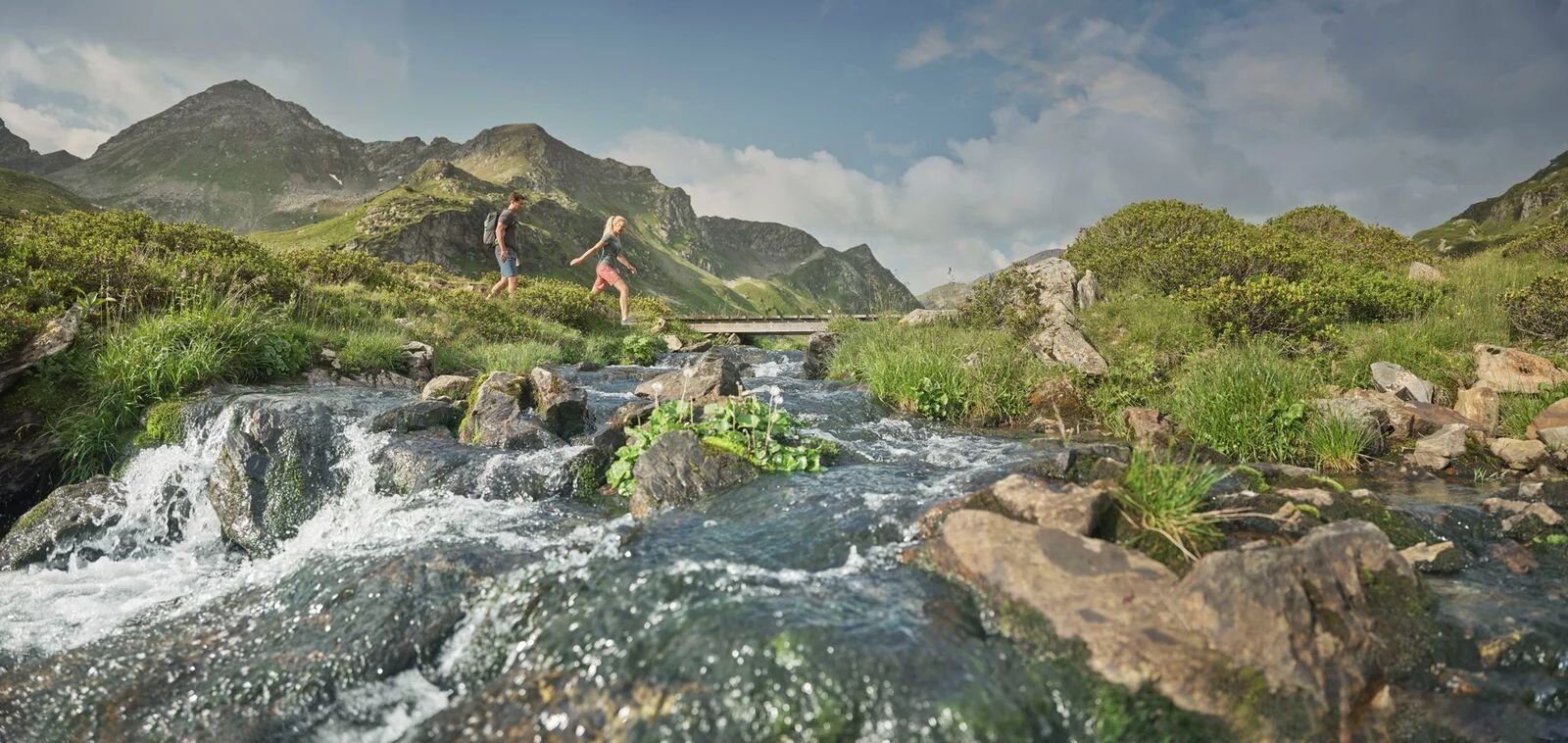 Two hikers cross a rocky stream in a mountain landscape.