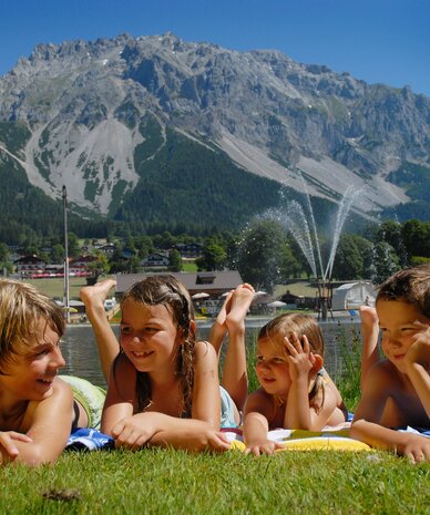 Four children lying on towels by lake with mountains