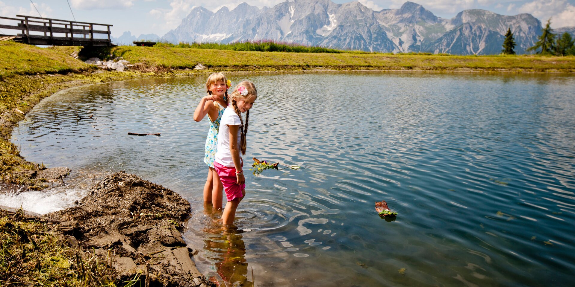 Two girls stand in a shallow alpine lake by a grassy bank.