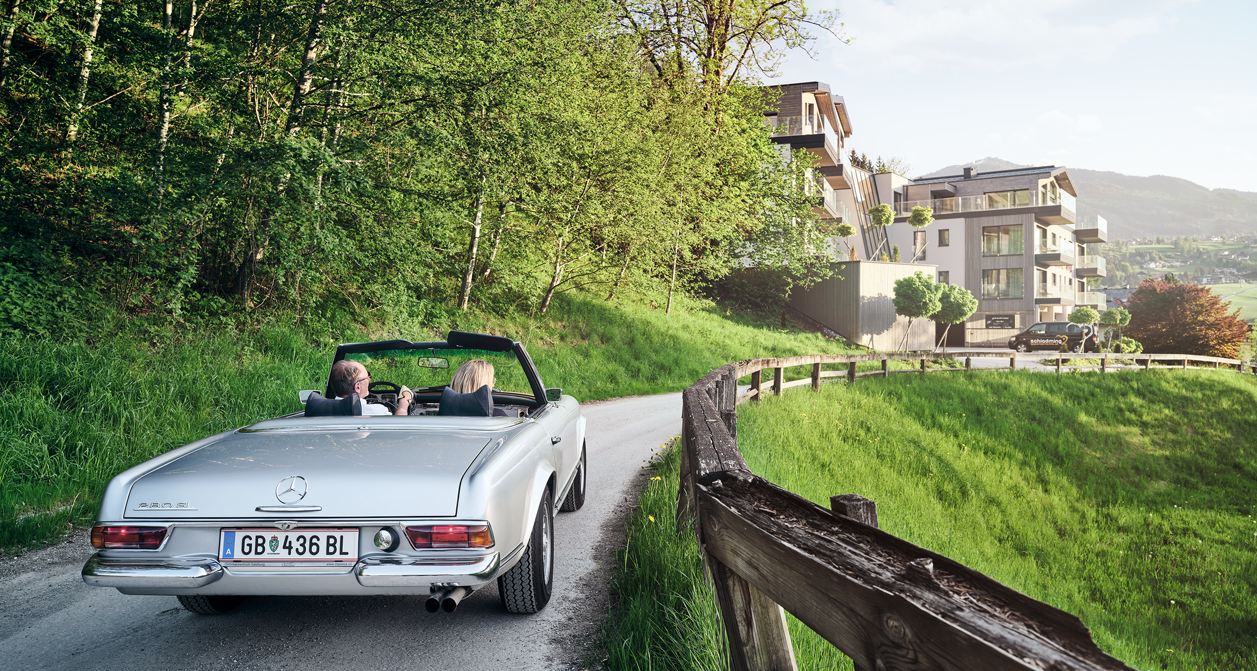 Silver convertible with two passengers on rural road near hillside apartments.