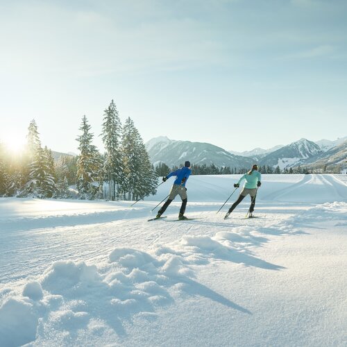 Two skiers glide across a snowy landscape toward forested mountains. | © Peter Burgstaller