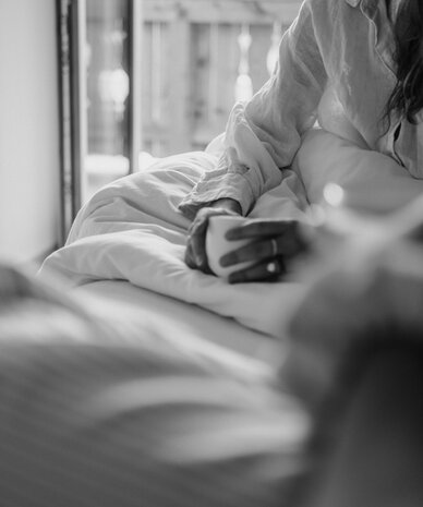 Person sitting up in bed holding a mug near the window.