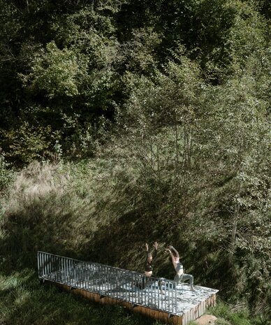 Two people practicing yoga on a raised platform in a forest clearing