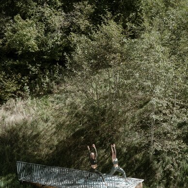 Two people practicing yoga on a wooden platform in a forest clearing