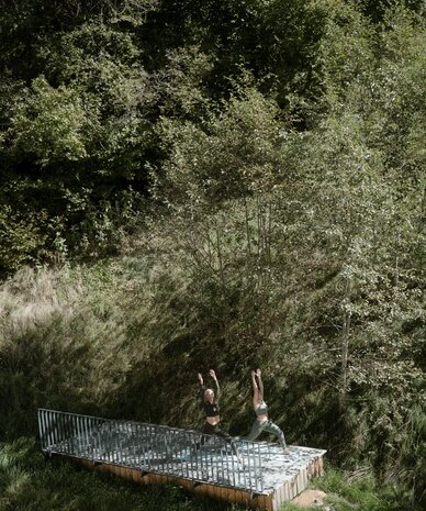 Two people practicing yoga on a wooden platform in a forest clearing