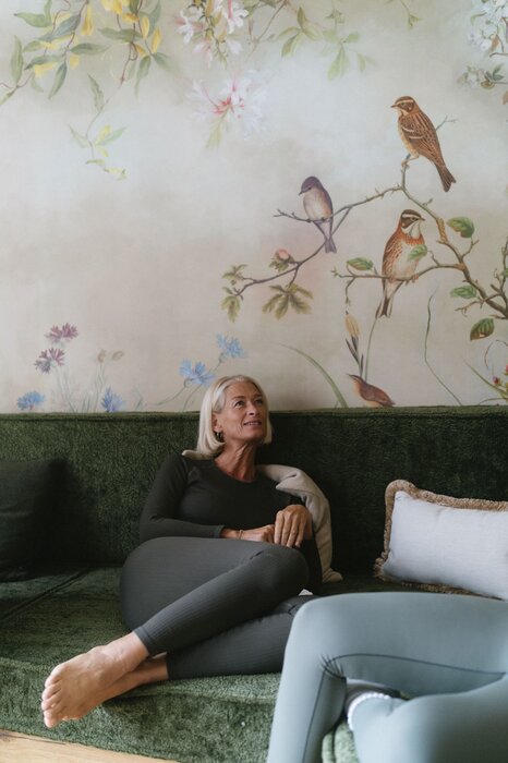 Older woman resting on green sofa with floral bird mural behind.