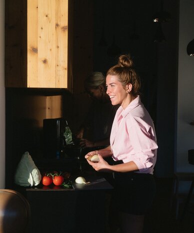 Woman in pink shirt chops vegetables in sunlit kitchen.